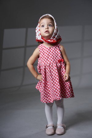 Foto de Retrato de una adorable niña rubia de pelo rizado en vestido a cuadros, con una bufanda, sobre fondo gris, plano de estudio - Imagen libre de derechos