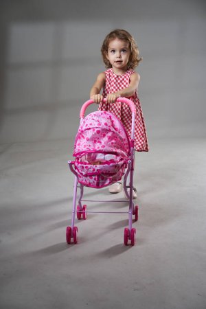 Photo for Portrait of an adorable blonde curly hair little girl in red plaid dress playing with a baby carriage toy on gray background, studio shot - Royalty Free Image