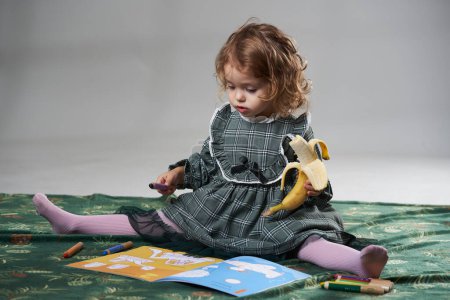 Foto de Retrato de una adorable niña rubia de pelo rizado comiendo un plátano y dibujando en un libro para colorear sobre fondo gris, filmado en estudio - Imagen libre de derechos