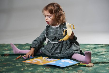Photo for Portrait of an adorable blonde curly hair little girl eating a banana and drawing on a coloring book on gray background, studio shot - Royalty Free Image