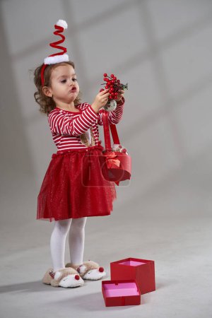 Téléchargez les photos : Portrait d'une adorable petite fille blonde aux cheveux bouclés en robe rouge et chapeau ouvrant cadeau de Noël sur fond gris, plan studio - en image libre de droit