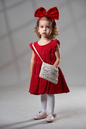 Téléchargez les photos : Portrait d'une adorable petite fille blonde aux cheveux bouclés en robe rouge et s'inclinant sur fond gris, plan studio - en image libre de droit