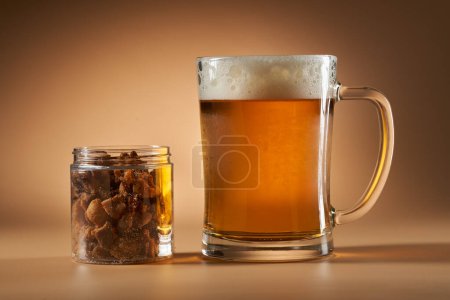 Photo for Pork greaves in a glass jar near a fresh cold beer in a mug, isolated on beige background - Royalty Free Image