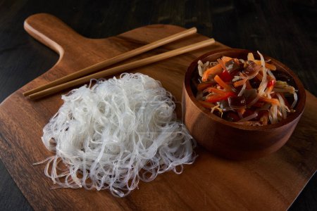 Téléchargez les photos : Hacher les légumes suey dans un bol et les nouilles en verre, une cuisine asiatique, sur une planche de bois - en image libre de droit