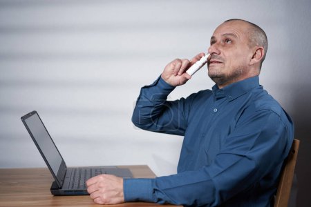 Photo for Mature businessman using nasal spray or inhaler at work, in front of his laptop - Royalty Free Image