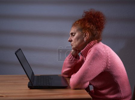 Foto de Pelirroja rizada freelancer mujer trabajando desde casa en su portátil, con una expresión de aburrimiento en su cara - Imagen libre de derechos