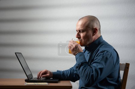 Photo for Businessman having a glass of fresh orange juice while working on the laptop at his desk - Royalty Free Image