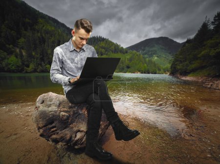 Foto de Conceptual image of a software developer or businessman working on laptop sitting on a stump by a beautiful lake in the mountains, mixed media - Imagen libre de derechos