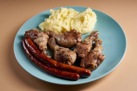 Téléchargez les photos : Jambes de poulet grillées et bâtonnets de saucisse avec plat d'accompagnement de pommes de terre en purée sur une assiette bleue sur fond beige - en image libre de droit
