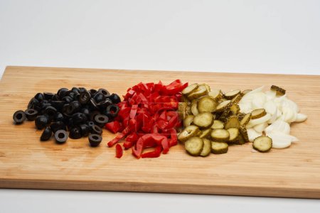 Foto de Mezcla de verduras en rodajas para una ensalada, con aceitunas negras, pimientos rojos en escabeche y pepinillos y cebolla fresca, sobre una tabla de madera - Imagen libre de derechos