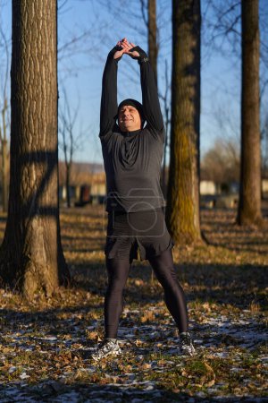 Téléchargez les photos : Coureur mature s'étirant avant de courir dans une journée froide et ensoleillée d'hiver - en image libre de droit