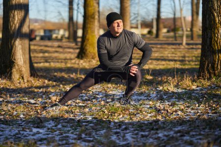 Téléchargez les photos : Mature coureur récréatif échauffement et étirement avant de courir dans une journée froide et ensoleillée d'hiver - en image libre de droit