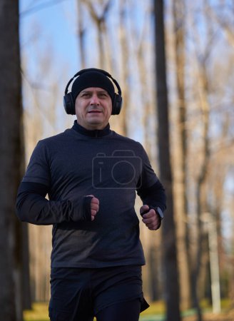 Photo for Mature runner with headphones in winter gear running in the park, listening to podcast or music - Royalty Free Image