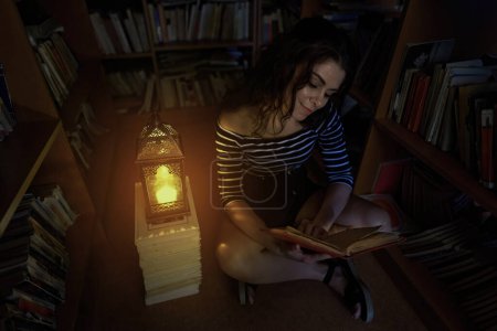 Téléchargez les photos : Jolie jeune bibliothécaire lisant la nuit à la bibliothèque, éclairée par une lanterne aux bougies - en image libre de droit