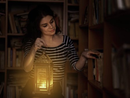 Foto de Atractivo joven bibliotecario leyendo por la noche en la biblioteca, iluminado por una linterna de vela - Imagen libre de derechos