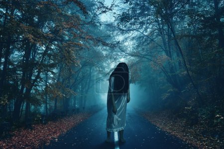 Photo for Zombie girl in night gown holding a soft rabbit toy walking barefoot in the middle of a road through a forest with mist, conceptual horror shot - Royalty Free Image
