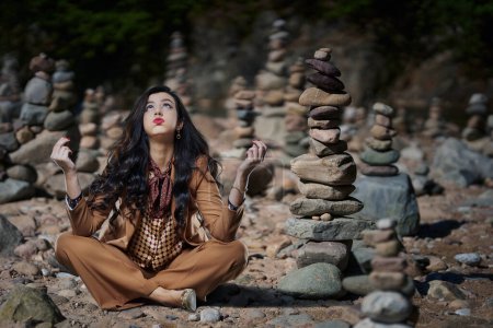 Photo for Businesswoman trying to chill after work stress, sitting among stacked zen stone pyramids on a beach - Royalty Free Image