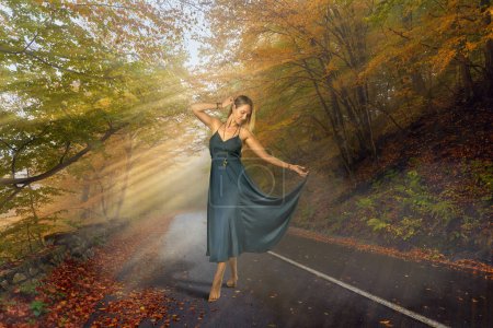 Photo for Beautiful tall blonde woman in blue dress and barefoot, dancing in the middle of the road in a misty morning with sun beams coming through forest - Royalty Free Image