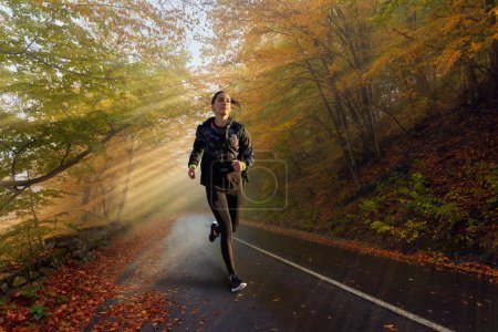 Photo for Female marathon runner training on a road through a misty forest in a cold autumn day with sun beams coming through - Royalty Free Image