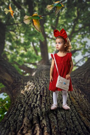 Fairytale little girl in an enchanted forest watching colorful birds fly by, standing on a huge tree