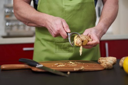 Téléchargez les photos : Homme cuisinier épluchage et nettoyage d'une grosse racine de gingembre sur une planche de bois - en image libre de droit