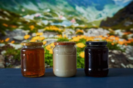 Téléchargez les photos : Différents types de miel dans des pots sur une planche en bois dans une belle vue sur la montagne avec des fleurs - en image libre de droit
