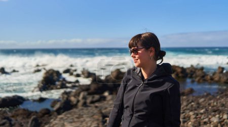 Téléchargez les photos : Belle jeune femme profitant d'une journée ensoleillée sur le rivage volcanique rocheux de Tenerife au bord de l'océan Atlantique - en image libre de droit