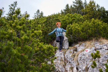 Téléchargez les photos : Femme alpiniste avec sac à dos escalade une montagne escarpée à travers les pinèdes - en image libre de droit