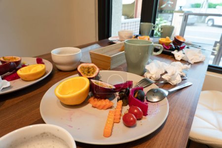 Foto de La mesa de desayuno está desordenada con comida sobrante, frutas, utensilios usados y platos. - Imagen libre de derechos