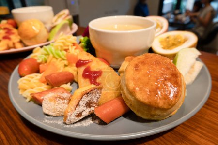 Foto de Un plato de diversos platos de desayuno, incluyendo salchichas, pescado frito, verduras, un bollo y una taza de sopa, con un fondo borroso de un entorno de comedor. - Imagen libre de derechos