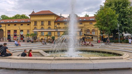 Foto de Szeged, Hungría - 30 de julio de 2022: Personas sentadas alrededor de la fuente de agua en la Plaza Dugonics, frente al Día de Verano del Edificio Universitario. - Imagen libre de derechos