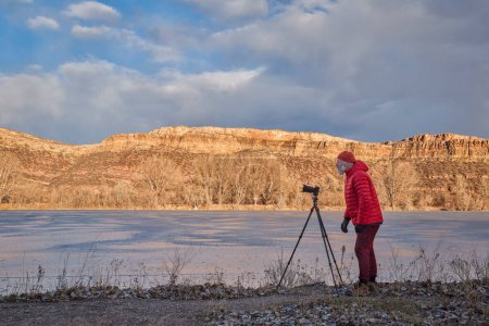 Photo for Senior male photographer with a camera on tripod on a shore of frozen lake in Colorado foothills - Royalty Free Image