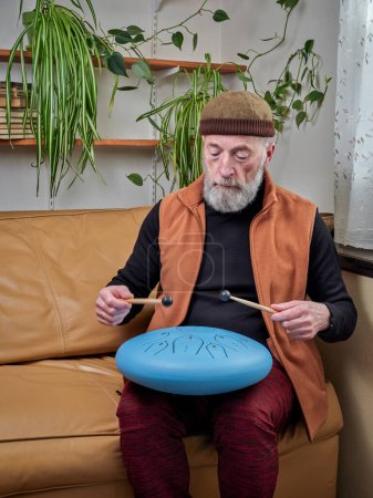Téléchargez les photos : Homme âgé concentré joue du tambour de langue en acier avec des maillets à la maison, style de vie et concept de passe-temps - en image libre de droit