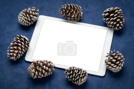 Téléchargez les photos : Maquette de tablette numérique avec écran isolé vierge (chemin de coupe inclus), pose plate avec cônes décoratifs en pin givré sur papier texturé bleu, thème vacances d'hiver - en image libre de droit