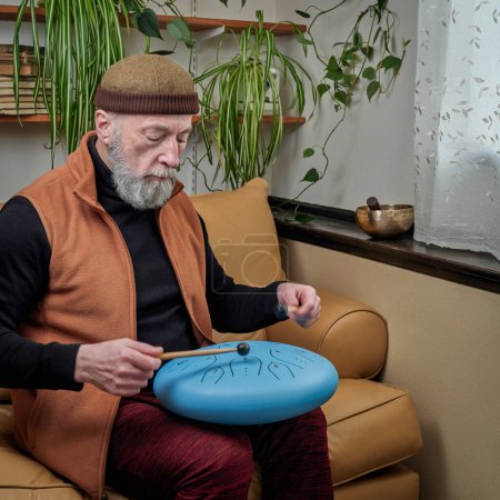 Téléchargez les photos : Homme âgé concentré joue du tambour de langue en acier avec des maillets à la maison, style de vie et concept de passe-temps - en image libre de droit