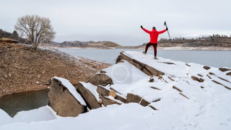 Foto de Happy male hiker with trekking poles on a rocky shore of a mountain lake at foothills of Rocky Mountains, Horsetooth Reservoir - a popular recreational area in northern Colorado in winter scenery - Imagen libre de derechos