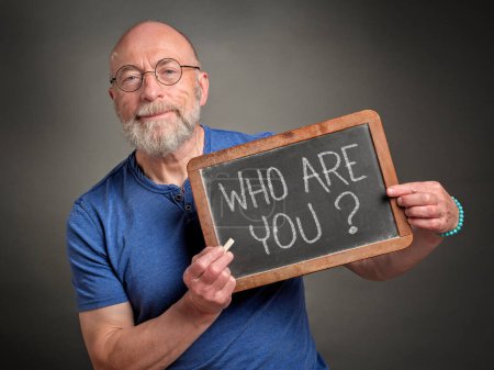Photo for Who are you? Senior man is sharing inspirational question handwritten on a blackboard. Philosophy, identity and personal development concept. - Royalty Free Image