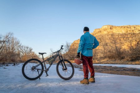 Foto de Senior male cyclist with a mountain bike contemplating winter sunset on a shore of Poudre River in northern Colorado - Imagen libre de derechos