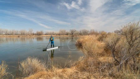 Téléchargez les photos : Senior male paddler is paddling stand up paddleboard on a calm lake in early spring scenery in Colorado - en image libre de droit