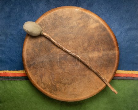 Photo for Handmade, native American style, shaman frame drum covered by goat skin with a beater against colorful abstract paper landscape - Royalty Free Image