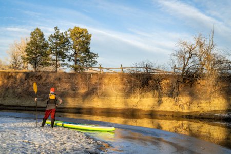Téléchargez les photos : Pagayeur mâle avec pédalier gonflable debout sur une rive glacée de la rivière Poudre à Fort Collins, Colorado - en image libre de droit