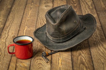 Téléchargez les photos : Chapeau outback en peau huilée avec tasse de thé en émail métallique sur une table en bois rustique - en image libre de droit