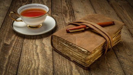Foto de Diario de cuero antiguo con una taza de té y pluma en una mesa de madera rústica - Imagen libre de derechos