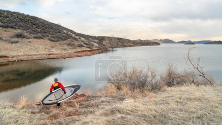 Téléchargez les photos : Pagayeur mâle en combinaison sèche et gilet de sauvetage porte sa planche à pagaie sur une rive du réservoir Horsetooth dans le nord du Colorado, pagaie d'hiver et concept d'entraînement - en image libre de droit