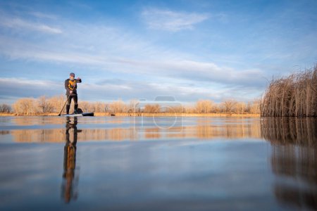 Photo for Senior paddler and his paddleboard on lake in winter or early spring in Colorado, frog perpective (partially submerged action camera) - Royalty Free Image