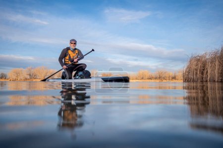 Téléchargez les photos : Pagayeur senior et sa planche à pagaie sur le lac en hiver ou au début du printemps dans le Colorado, perspective grenouille (caméra d'action partiellement submergée) - en image libre de droit