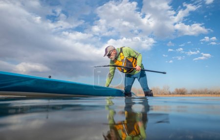 Téléchargez les photos : Pagayeur senior et sa planche à pagaie sur le lac en hiver ou au début du printemps dans le Colorado, perspective grenouille (caméra d'action partiellement submergée) - en image libre de droit