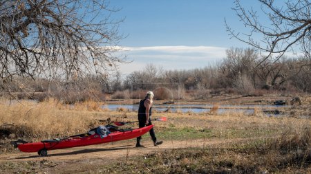 Foto de Evans, CO, USA - 1 de abril de 2023: Un kayak más viejo y masculino está transportando su kayak de mar en un carro para remar a principios de primavera en el río Platte Sur en Colorado. - Imagen libre de derechos