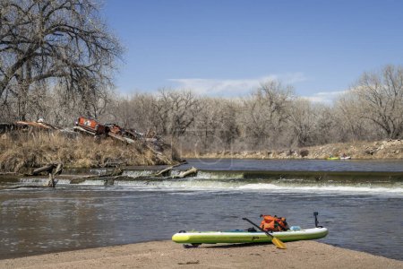 Téléchargez les photos : Planche à pagaie gonflable debout avec une pagaie, un sac sec et une caméra d'action sur une barre de sable en dessous du barrage de dérivation, pagaie au début du printemps sur la rivière South Platte dans le Colorado. - en image libre de droit