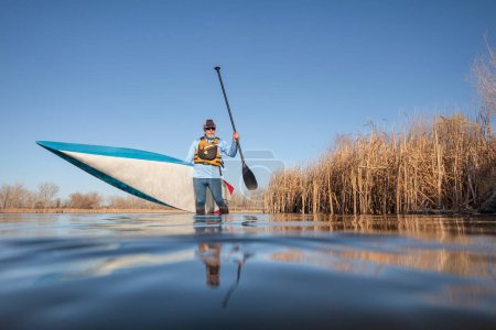 Téléchargez les photos : Senior mâle pagayeur lance un stand up paddleboard sur un lac calme au début du printemps, perspective grenouille d'une caméra d'action au niveau de l'eau - en image libre de droit